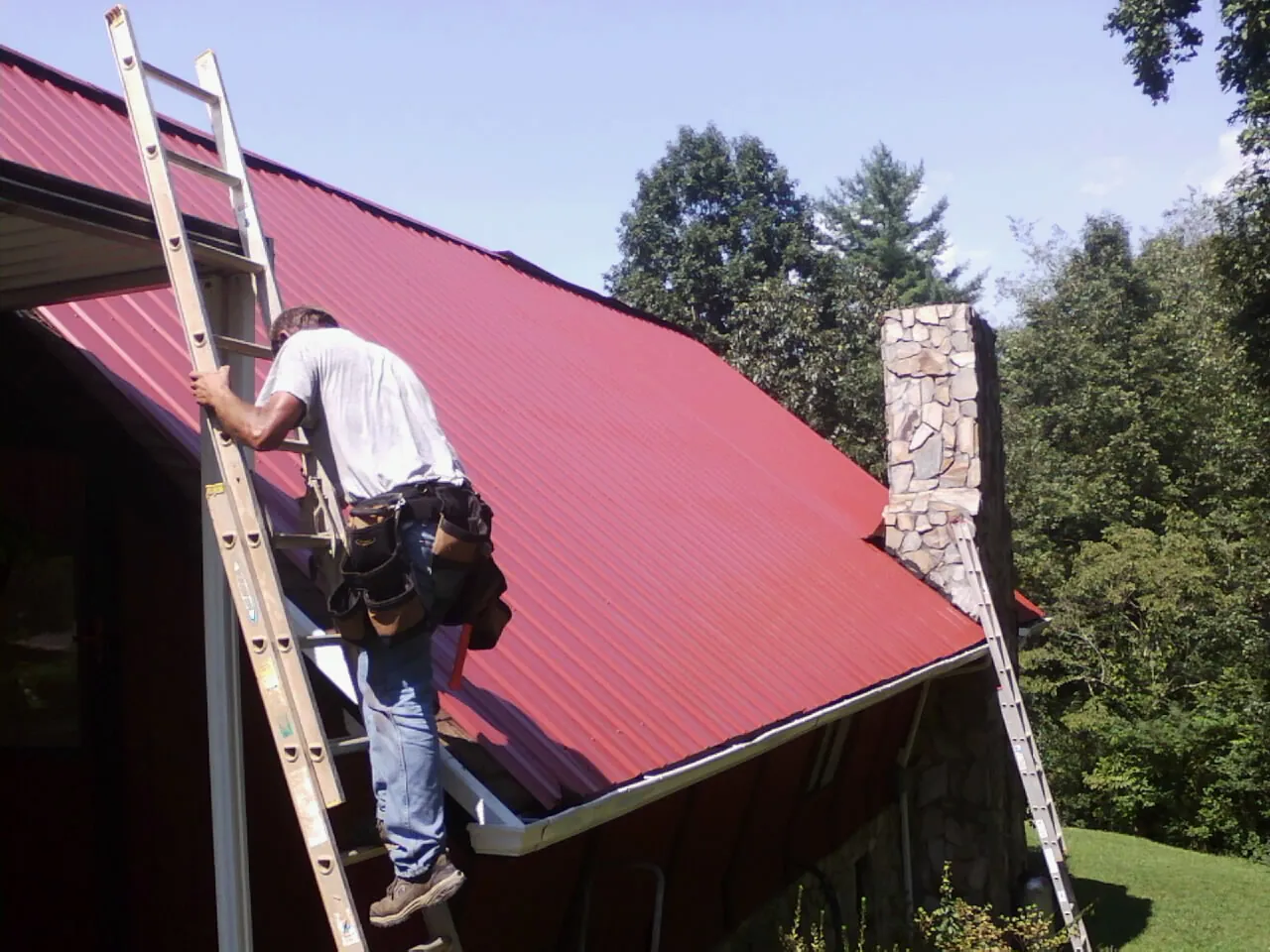 Roofer on a ladder beside a red metal roof and stone chimney — Soffit & Fascia Repair by Roofing Brothers in Lovejoy